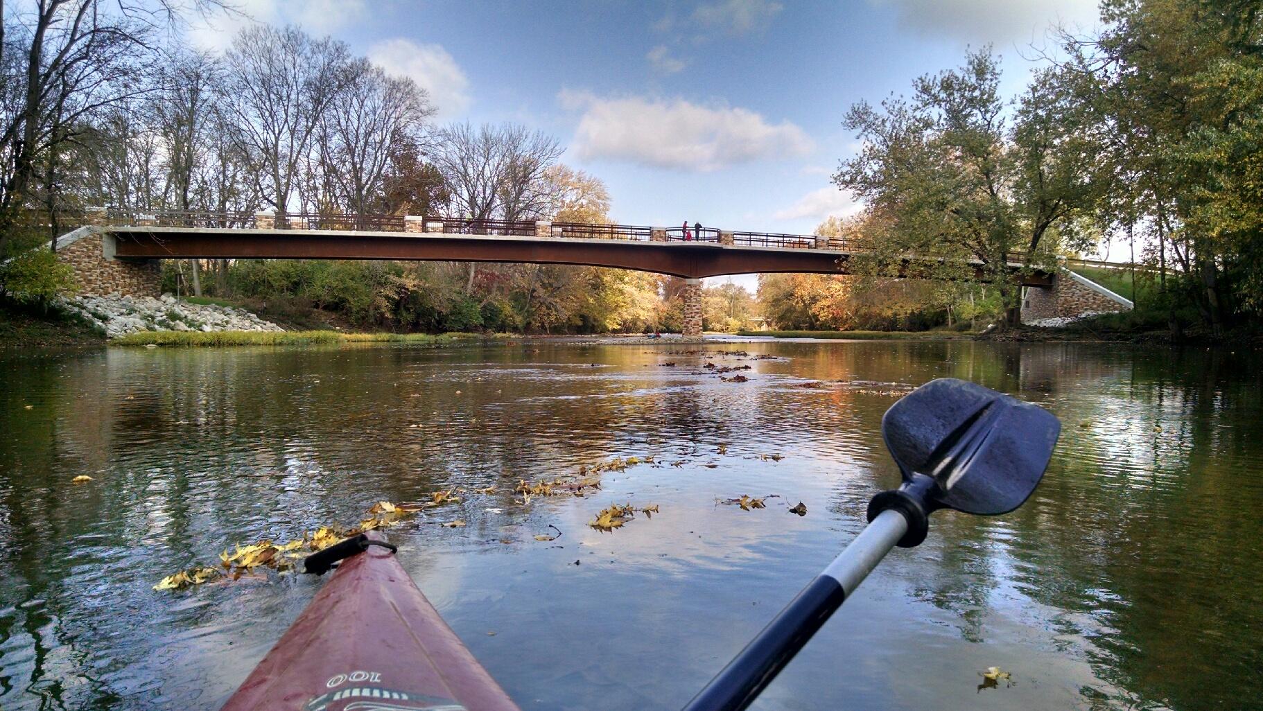 A kayaker paddles on the Great Miami River with a bridge seen ahead. Trees line the banks
